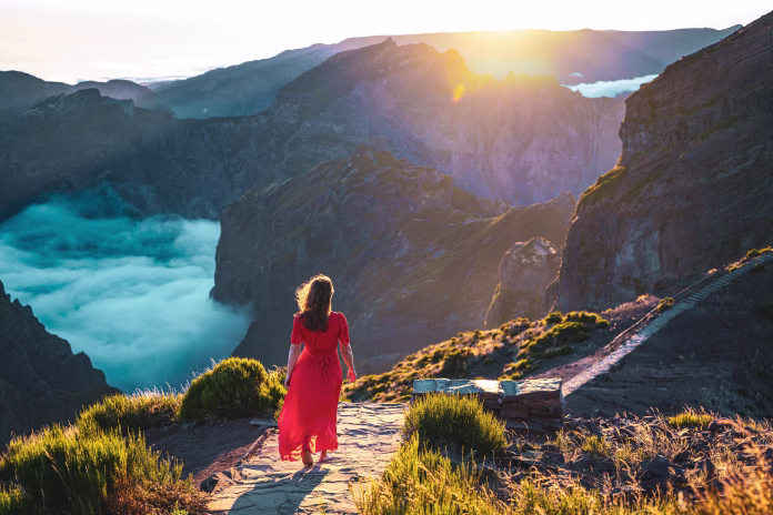 Description: Beautiful woman in red dress walking barefoot on a very scenic hiking trail in the evening sun on Pico do Ariero. Verade do Pico Ruivo, Madeira Island, Portugal, Europe.