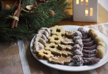 Ne nasedajte! Piškoti, ki jih imamo za ‘domače’, skrivajo šokantne sestavine Gingerbread, Linzer and other types of Christmas cookies on a white plate, a festive tablecloth, an Advent wreath and a white decorative house with a burning candle in the background. Selective focus