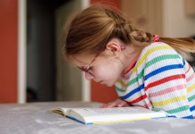 Nič več liberaliziranega trga dragih učnih gradiv: sprejet Zakon o potrjevanju učnih gradiv Little happy girl with eyeglasses reading book at home. Elementary school child reads, making homework. Education concept.