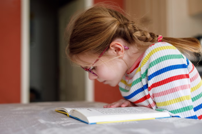 Little happy girl with eyeglasses reading book at home. Elementary school child reads, making homework. Education concept.