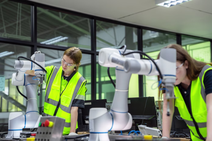 Two robots are being worked on by two people. One of the people is wearing a yellow vest