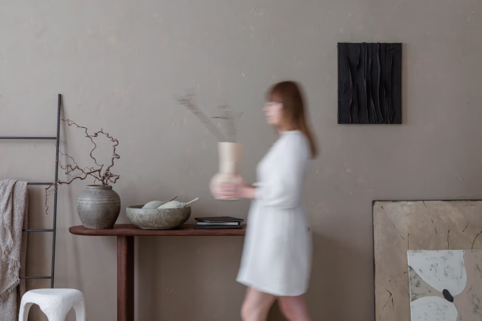 Aesthetic composition of warm living room interior with mock up poster frame, woman with vase,  wooden consola, gray bowl, black ladder, beige wall and personal accessories. Home decor. Template.