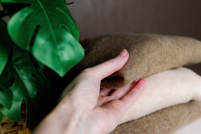 Caucasian woman takes with one hand a coffee-colored cashmere sweater from a white nightstand with a green monstera flower, extreme close-up side view. Conceptual women's clothing, new collections, natural items, fashion industry.