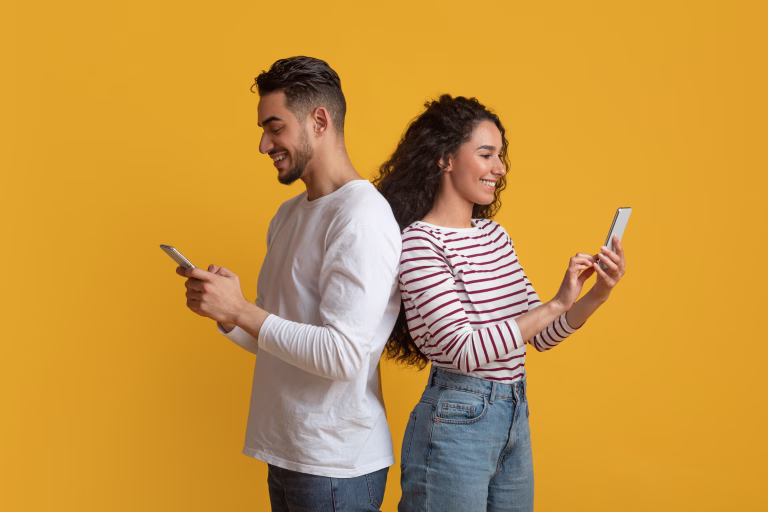 Gadget Addiction. Young Arab Couple Holding Smartphones And Standing Back To Back, Happy Middle Eastern Man And Woman Busy With Cellphones While Standing Together On Yellow Background, Copy Space