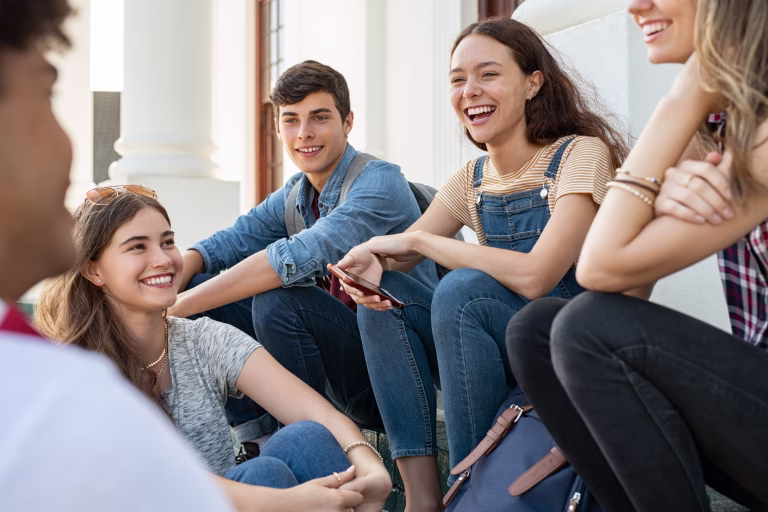 Group of happy young friends sitting in college campus and talking. Cheerful group of  smiling girls and guys feeling relaxed after university exam. Excited millenials laughing and having fun outdoor.