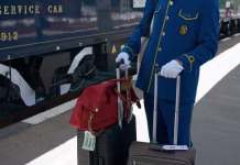 Kako doživeti čar potovanja z Orient Expressom v letu 2026 (tudi s študentskim proračunom) June 03,2025 - Ruse, Bulgaria: White-gloved Venice Simplon Orient Express steward in blue and gold uniform wheels guest luggage beside a navy service car, showcasing Art Deco elegance and premium European rail hospitality.