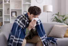 Zakaj nekateri umrejo zaradi sicer nenevarnih okužb? Man sitting on couch wrapped in a blue blanket, sneezing into tissue. Scene depicts illness, cold, flu symptoms, and seeking comfort at home. Expression of discomfort and need for care.