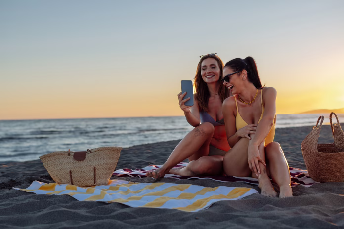 Two women laughing on a beach at sunset while sitting on a towel next to each other and looking at something funny on a smartphone.