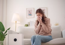 Tako boste zmanjšali težave z alergijami v vašem domu Young Asian woman sits on a couch, sneezing and holding a tissue to her nose while an air purifier operates nearby to improve air quality.