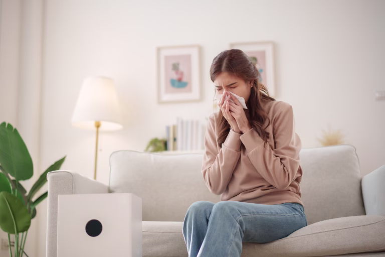Young Asian woman sits on a couch, sneezing and holding a tissue to her nose while an air purifier operates nearby to improve air quality.