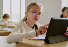 Napaka pri vzgoji nadarjenih otrok, ki jo je odkrila znanost Young girl wearing glasses sitting at wooden desk using tablet in classroom surrounded by fellow students in bright learning environment