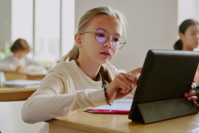 young-girl-wearing-glasses-sitting-at-wooden-desk-using-tablet-in-classroom-surrounded-by-fellow-students-in-bright-learning-environment-stockpack-istock Young girl wearing glasses sitting at wooden desk using tablet in classroom surrounded by fellow students in bright learning environment