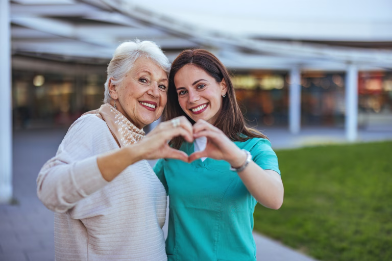 Elderly woman and her caregiver happily form a heart shape with their hands, symbolizing care and companionship in an outdoor setting.