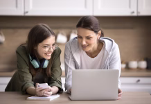 5 vprašanj, ki jih naj starši vprašajo po informativcih Pretty teen girl holding smartphone talking to mature mother helps her with on-line school assignment using laptop, prepare task standing together in kitchen. Education using modern tech, applications