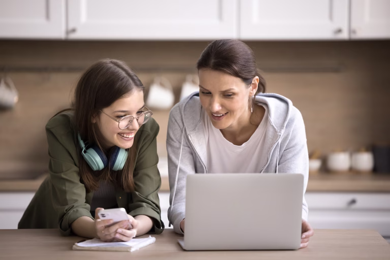 Pretty teen girl holding smartphone talking to mature mother helps her with on-line school assignment using laptop, prepare task standing together in kitchen. Education using modern tech, applications