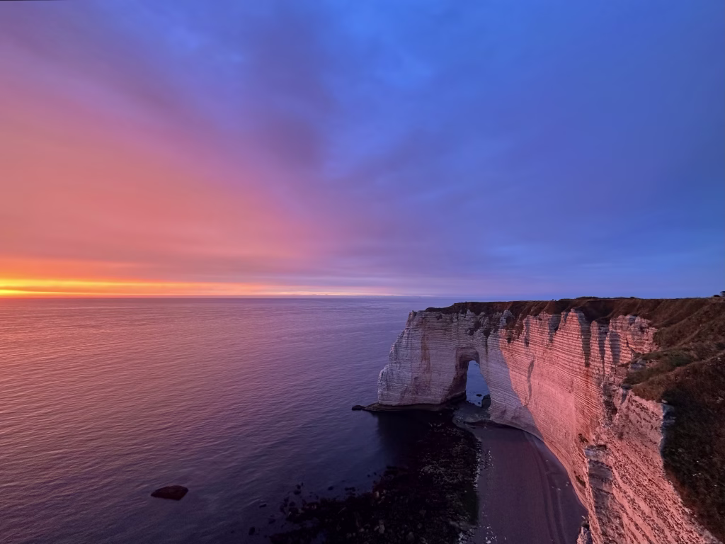 Purple and orange hues illuminate the sky over the Cliffs of Étretat, located in Normandy, France, alongside the English Channel. The iconic limestone cliffs, featuring natural arches and rugged formations, stand prominently against the tranquil waters. The scene, captured during sunset, emphasizes the striking contrast between the vibrant sky and the pale white of the cliffs, highlighting the natural beauty and dramatic landscape of this coastal region.