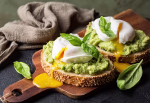 Ta trik za poširana jajca je obnorel internet: s tem bo zajtrk še bolj popoln Smashed avocado sandwich with boiled poached egg with flowing yolk and basil leaves. Dark green background.