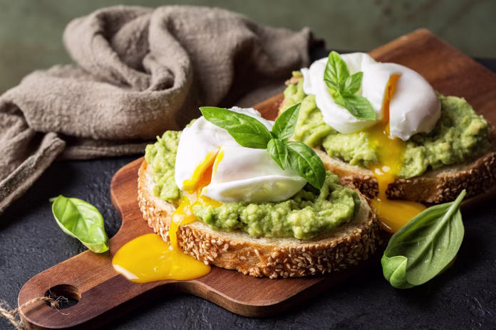 Smashed avocado sandwich with boiled poached egg with flowing yolk and basil leaves. Dark green background.