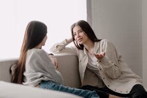Two millennial woman talking and gesturing, sitting on comfort couch at home. Young female spending weekend together, having pleasant conversation, sharing secrets, enjoying company