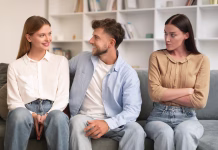 Partner je prijatelj z bivšo: vzrok za preplah ali nič posebnega? Young man hugs his girlfriend on couch while another woman looks with jealousy crossing hands, sitting together on sofa in modern living room. Infidelity, complicated romantic triangle