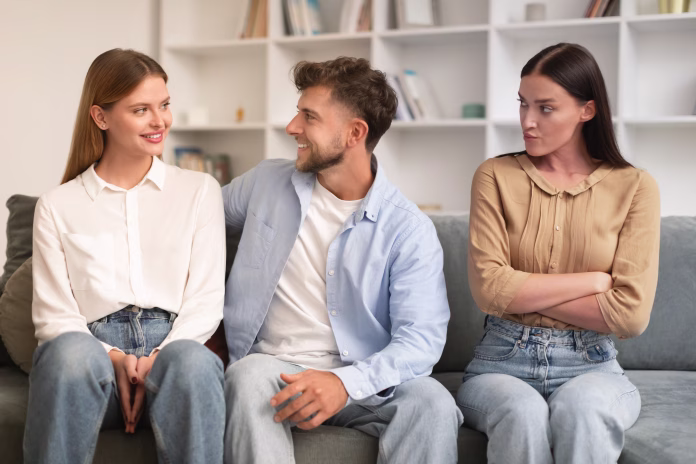 Young man hugs his girlfriend on couch while another woman looks with jealousy crossing hands, sitting together on sofa in modern living room. Infidelity, complicated romantic triangle