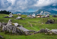 Velika planina: le uro iz Ljubljane do enega najlepših razgledov v Sloveniji A wide panoramic view of a traditional shepherd village with unique wooden huts scattered across green rolling hills under a dramatic sky in Velika Planina, Slovenia.