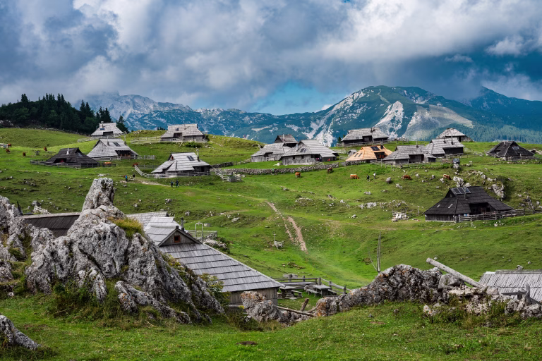 A wide panoramic view of a traditional shepherd village with unique wooden huts scattered across green rolling hills under a dramatic sky in Velika Planina, Slovenia.