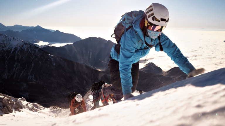 Climber in a blue jacket and helmet ascends a snowy peak, flanked by two companions, symbolizing teamwork and adventure in a breathtaking mountain landscape.