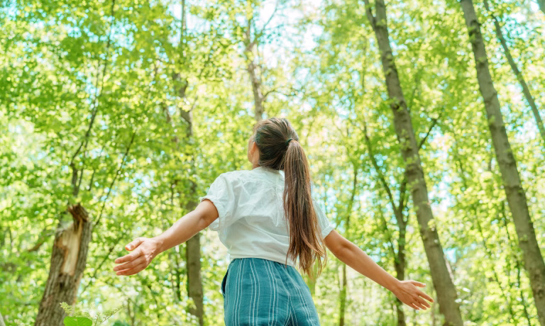 Free woman breathing clean air in nature forest. Happy girl from the back with open arms in happiness. Fresh outdoor woods, wellness healthy lifestyle concept.