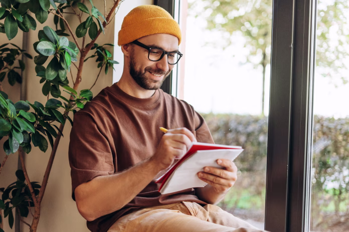 Portrait of smiling, happy, bearded man wearing stylish eyeglasses and yellow hat taking notes, writer having paperwork, sitting at home. Education concept