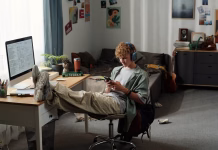 Pravilo 5 sekund: tako premagate prokrastinacijo Teenage Caucasian boy sitting in messy bedroom wearing headphones, leaning back with feet on desk, intensely using smartphone while ignoring computer and study materials nearby