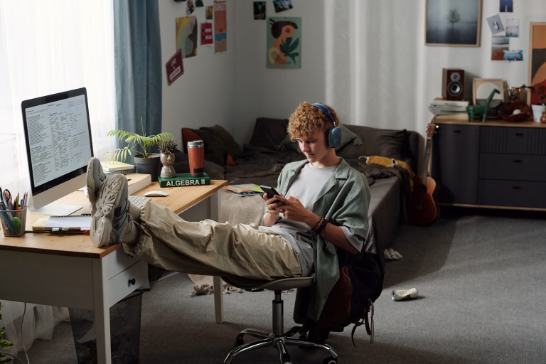 Teenage Caucasian boy sitting in messy bedroom wearing headphones, leaning back with feet on desk, intensely using smartphone while ignoring computer and study materials nearby