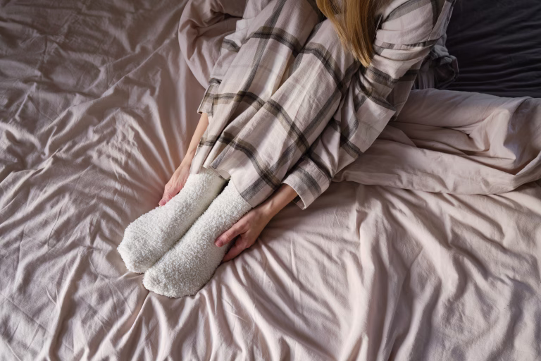 The girl lies in bed in pajamas and warm socks. Women's legs in cozy white fluffy socks in bed. View from above. Warm evenings at home