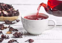 Ali so aromatizirani čaji nezdravi? White cup of healthy hibiscus tea pouring from the teapot with dried hibiscus flowers on white wooden background, winter hot drink concept for cold and flu