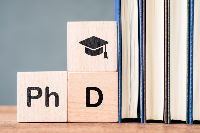Wooden blocks displaying PhD text and graduation cap symbol placed beside stacked books, representing advanced academic study, higher education goals, research degree, and scholarly achievement