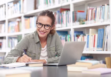 5 korakov za pregled literature: le tako bo zaključno delo zaokrožena celota Young female university student studying in a library, sitting at a desk with a computer and writing in a notebook. Concept of education, knowledge, learning, academic success, and exam preparation.