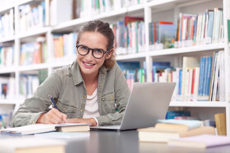 Young female university student studying in a library, sitting at a desk with a computer and writing in a notebook. Concept of education, knowledge, learning, academic success, and exam preparation.