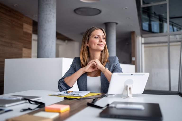 Businesswoman sitting at a modern office desk, smiling and gazing thoughtfully into the distance while contemplating future business plans and aspirations for success