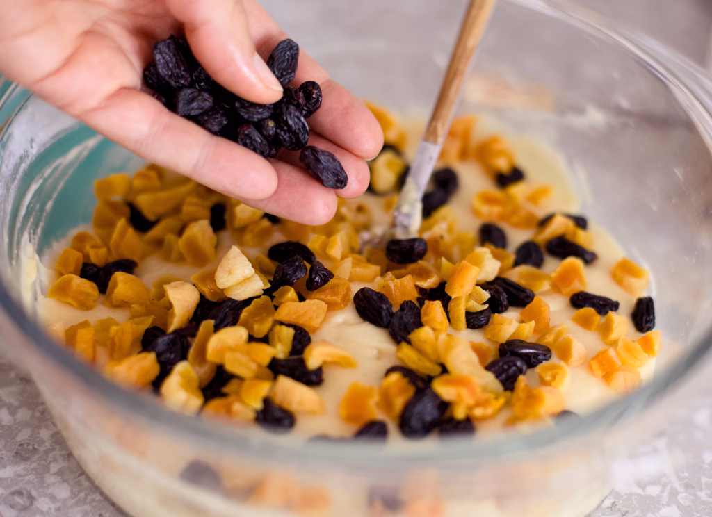 Close-up shot of a woman hand adding piece of dried fruits to dough that is in a glass bowl on a kitchen table