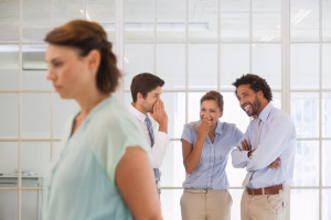 Colleagues gossiping with sad young businesswoman in foreground at a bright office