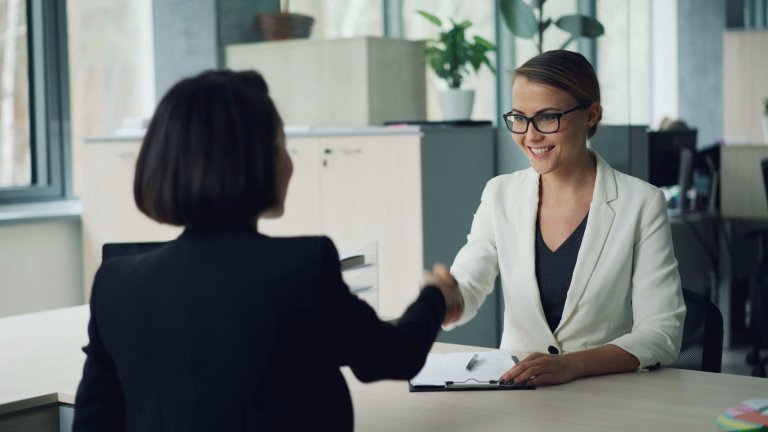 Good-looking young lady from human resources department is interviewing a female professional shaking hands then asking questions and writing. Employment and successful youth concept.