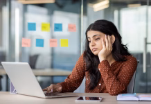 Kaj narediti, ko vam v službi postane dolgčas? Latin American businesswoman appears stressed at work, gazing at laptop screen in modern office. Creative ideas on sticky notes surround her, with smartphone by her side, reflecting focus.