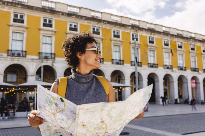 Smiling tourist is holding a map and wearing a yellow backpack while visiting the beautiful city of lisbon in portugal during summer vacations