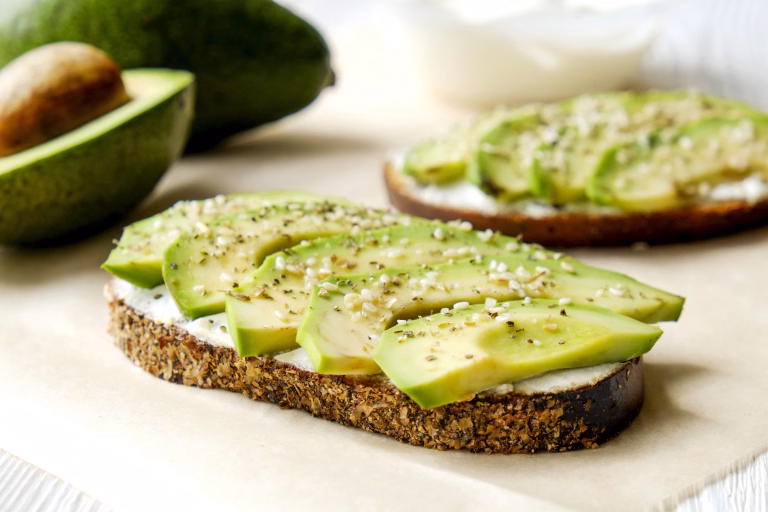 Vegan sandwich, rye bread toast, avocado slices, vegenaise sauce & raw arugula. Toasted sourdough, eggless mayonnaise, wooden table, whole & halved. National avocado day concept. Close up, background.