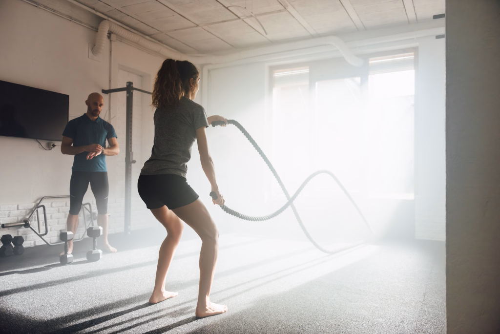 Anonymous barefoot female athlete looking away while standing with bent legs and exercising with battle ropes with help of male instructor in back lit sunbeam from gym glass window