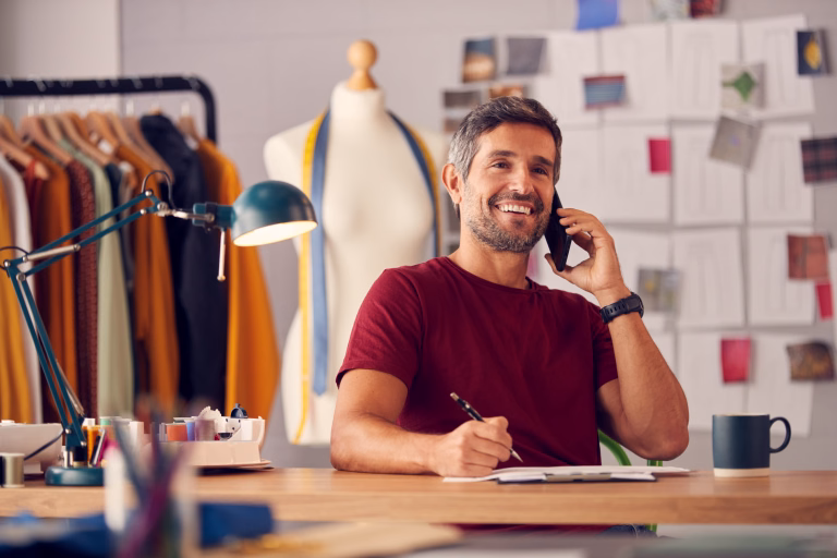 Mature Male Fashion Designer In Studio Working On Sketches Or Documents At Desk Making Phone Call