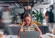 15 nasvetov za izboljšanje koncentracije in fokusa Young adult woman feeling stressed and focused, working on a laptop in a bustling modern coworking space with motion blurred people