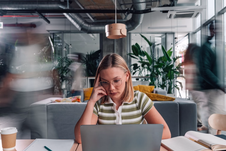 Young adult woman feeling stressed and focused, working on a laptop in a bustling modern coworking space with motion blurred people