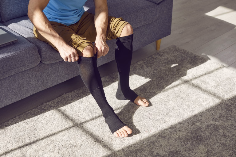 Young man wears compression stockings due to thrombosis or vein swelling. Unrecognizable man sitting on sofa and putting elastic medical anti varicose or anti thrombotic sleeve socks on his tired legs