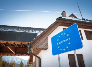 Konec poceni nakupov: Od 1. julija velika sprememba pri nakupih – tako višje bodo cene A blue roadside panel with the 12 stars of the European Union and the word 'Slovenija' marks the entrance to Slovenia on the route from Austria. The sign stands near buildings of a former checkpoint in a mountain setting, illustrating the Schengen system where routine border controls have been removed and movement is simplified for travelers. This clear, close composition is useful for stories on EU membership, mobility, tourism flows, cross-border commuting, transport policy, and the visual language of European road signage at internal borders.
